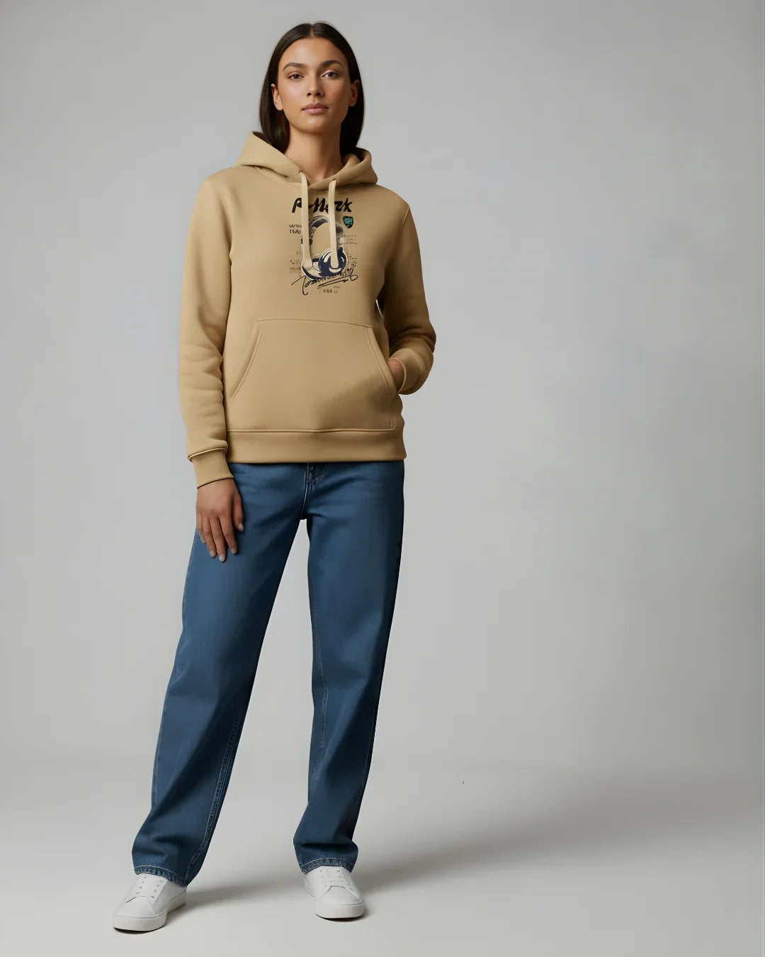 Woman wearing beige graphic hoodie, blue jeans, and white sneakers on plain background