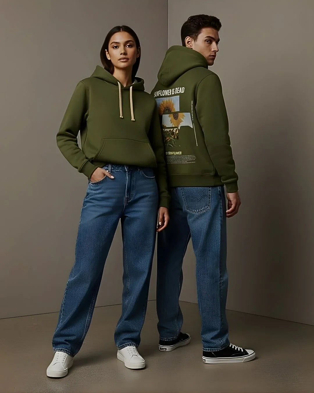 Two models in olive green hoodies and blue jeans pose in a minimalist studio setting.
