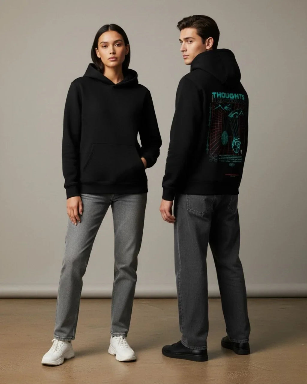 Two models wear black hoodies and grey jeans against a plain indoor backdrop.