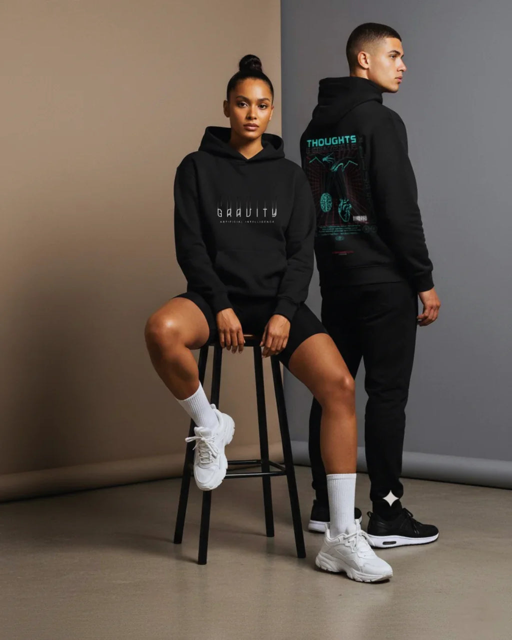 Two models wear black Gravity AI hoodies, one seated and one standing in a minimalist studio.