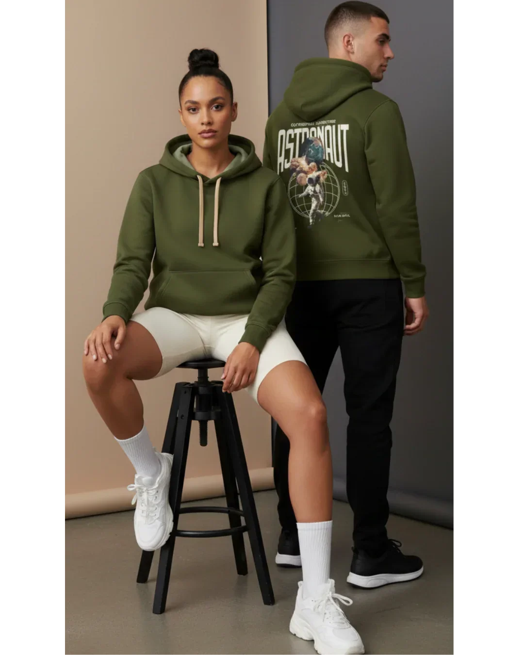 Two models in olive green astronaut graphic hoodies, casual athleisure studio setting